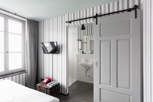 Una habitación de hotel moderna con una cama, un teléfono rojo, una maleta, un televisor montado en la pared y un baño con lavabo y espejo.