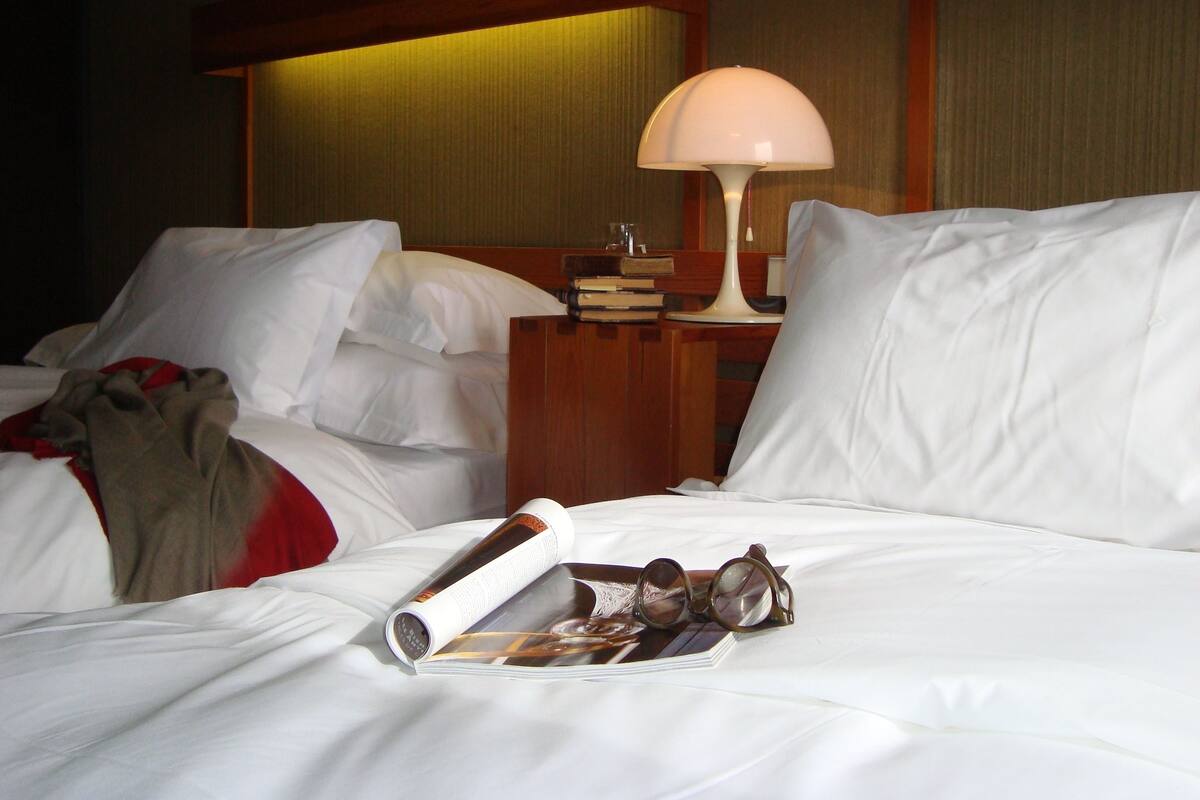 Una habitación de hotel con una cama, una mesita de noche con una lámpara y una revista con vasos encima.