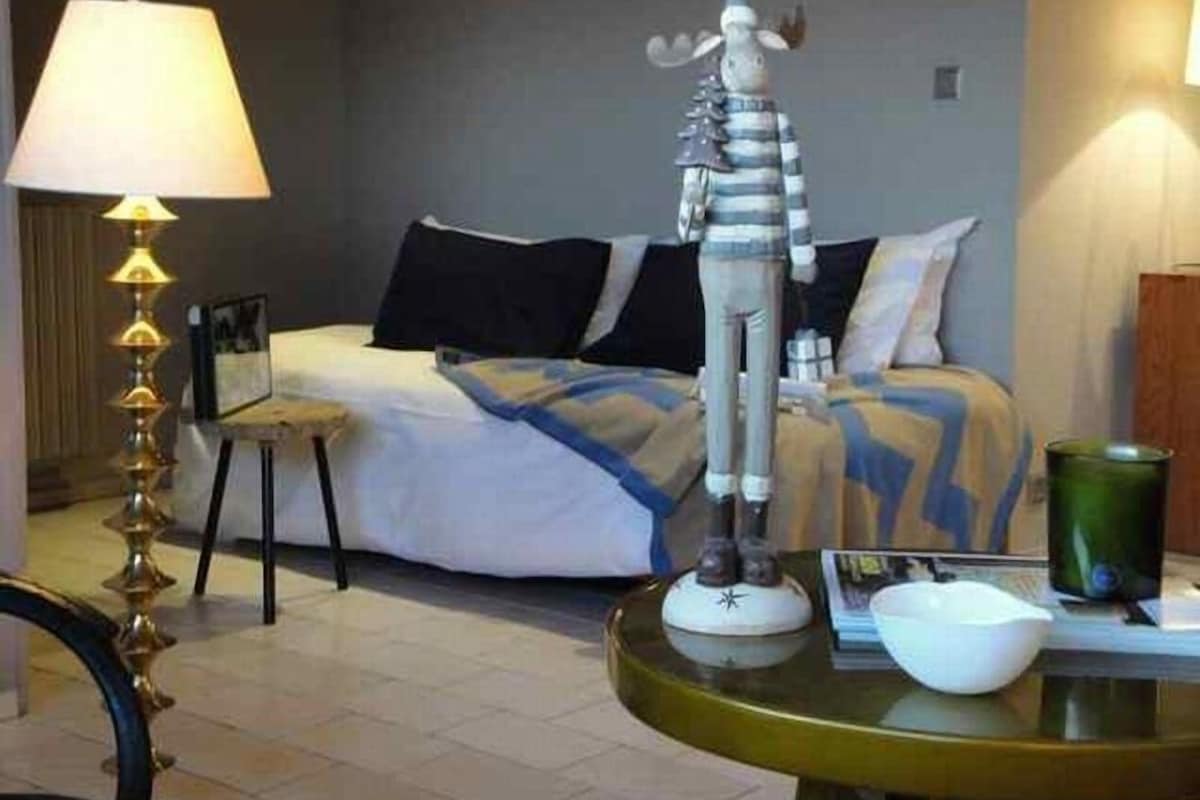 Habitación de hotel con una cama, una lámpara de pie, una mesita con un tazón y una taza, y un adorno que parece un alce.