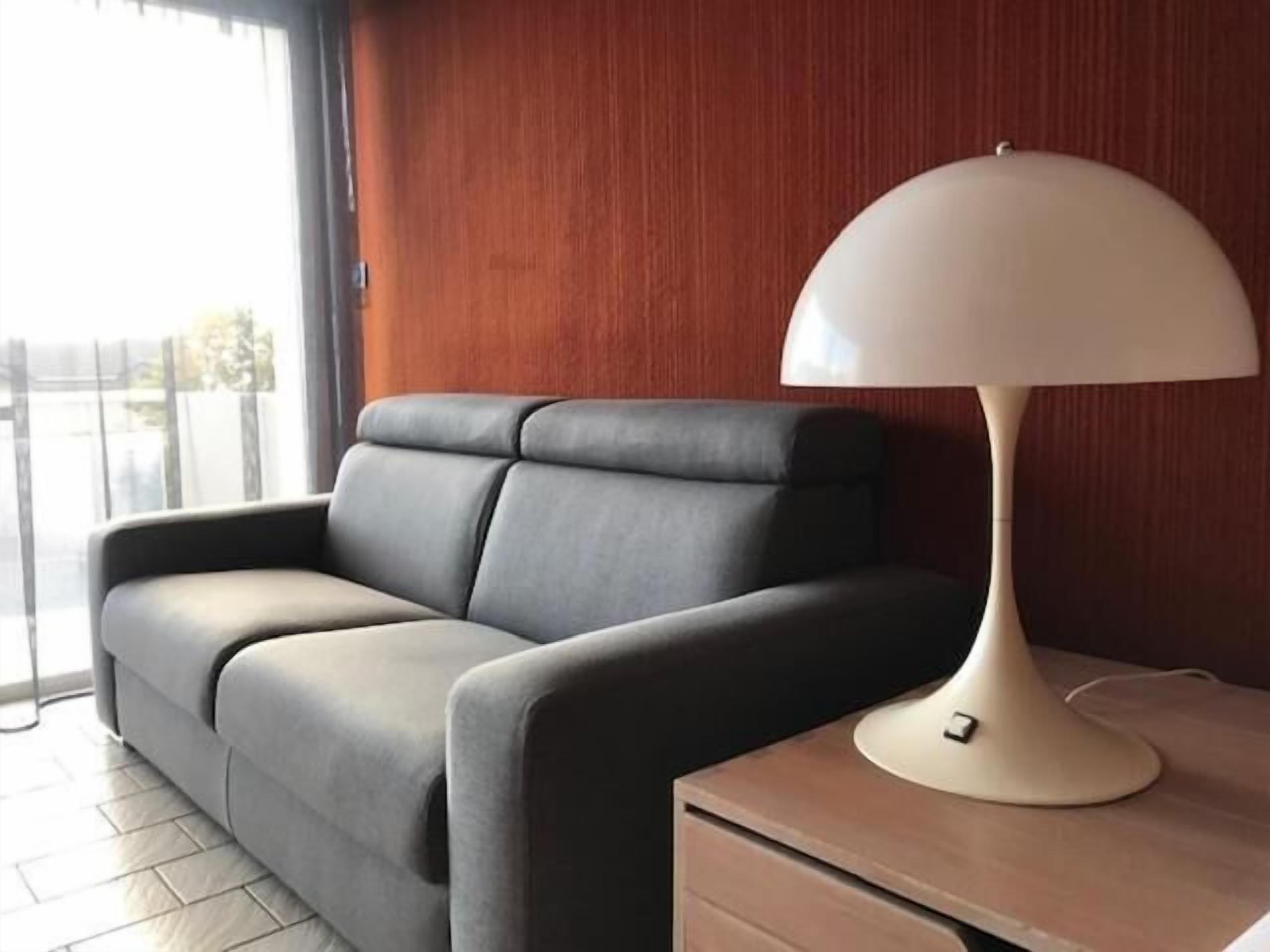 Een moderne kamer met een grijze bank, een houten nachtkastje en een witte lamp.