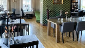 Un comedor espacioso con techos altos, detalles ornamentales en el techo y una araña. Las mesas están dispuestas con sillas, y se ve una silla tapizada de color verde.