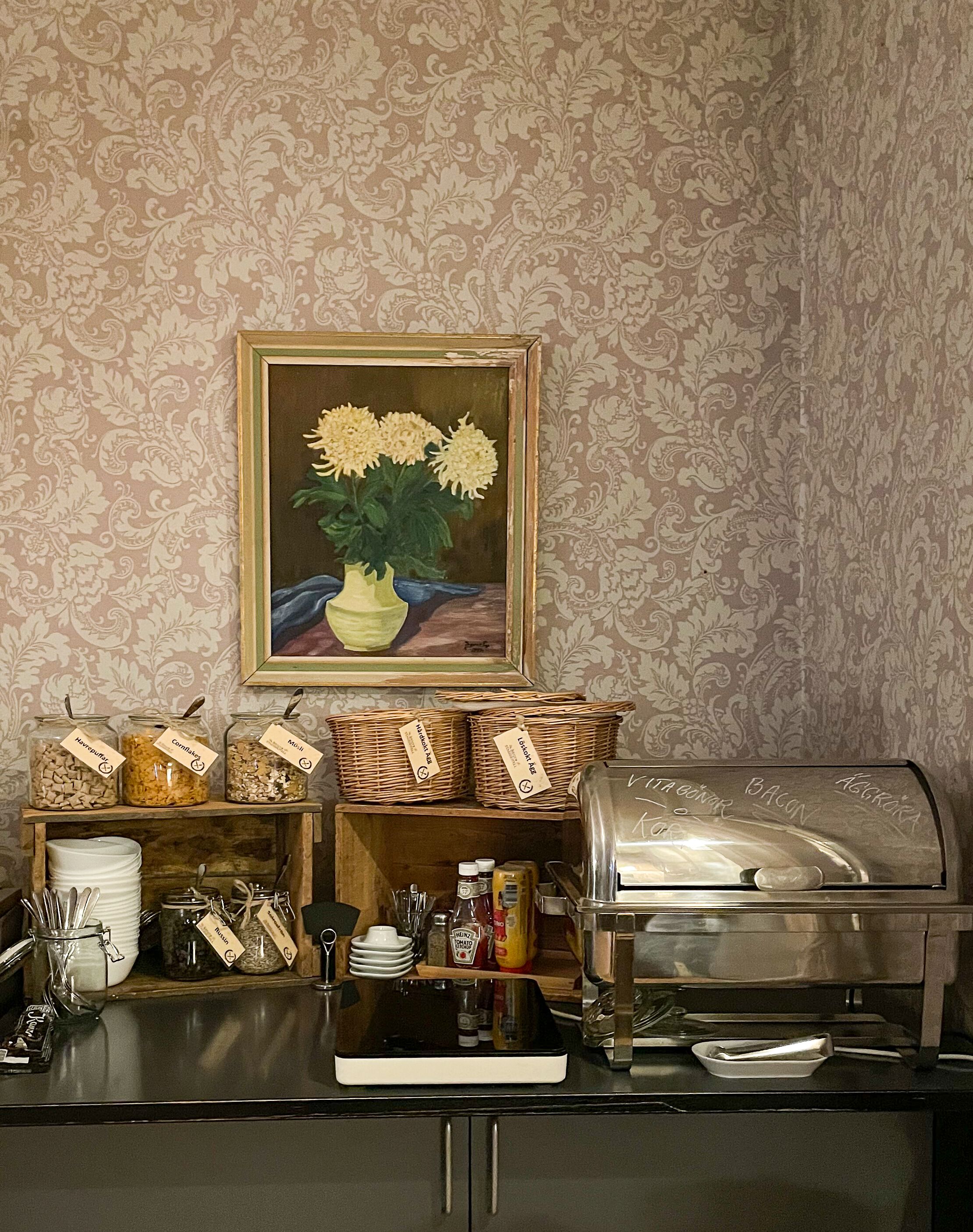 Un buffet de petit-déjeuner avec des pots d'étalement clairement étiquetés, un coin café et un tableau représentant des fleurs.