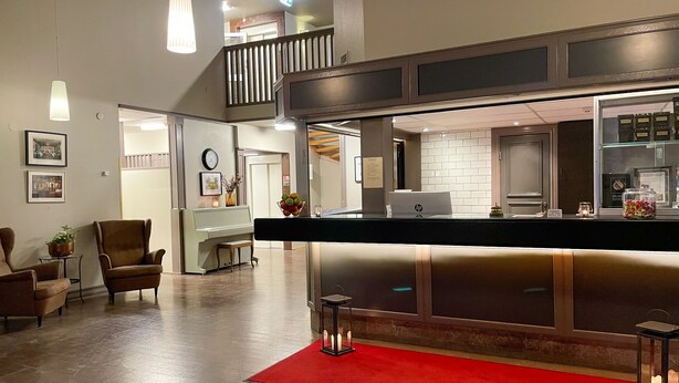 Un moderno hall de hotel con mostrador de recepción, un piano, zona de estar y una escalera que conduce a un nivel superior.