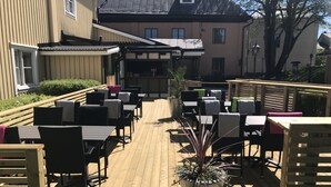 Un patio al aire libre con mesas y sillas de madera, una planta en maceta y un edificio al fondo.