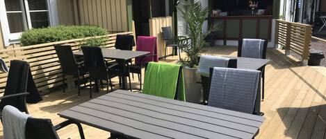 Un patio al aire libre con cubierta de madera, mesas y sillas de metal, y una pequeña zona de bar.