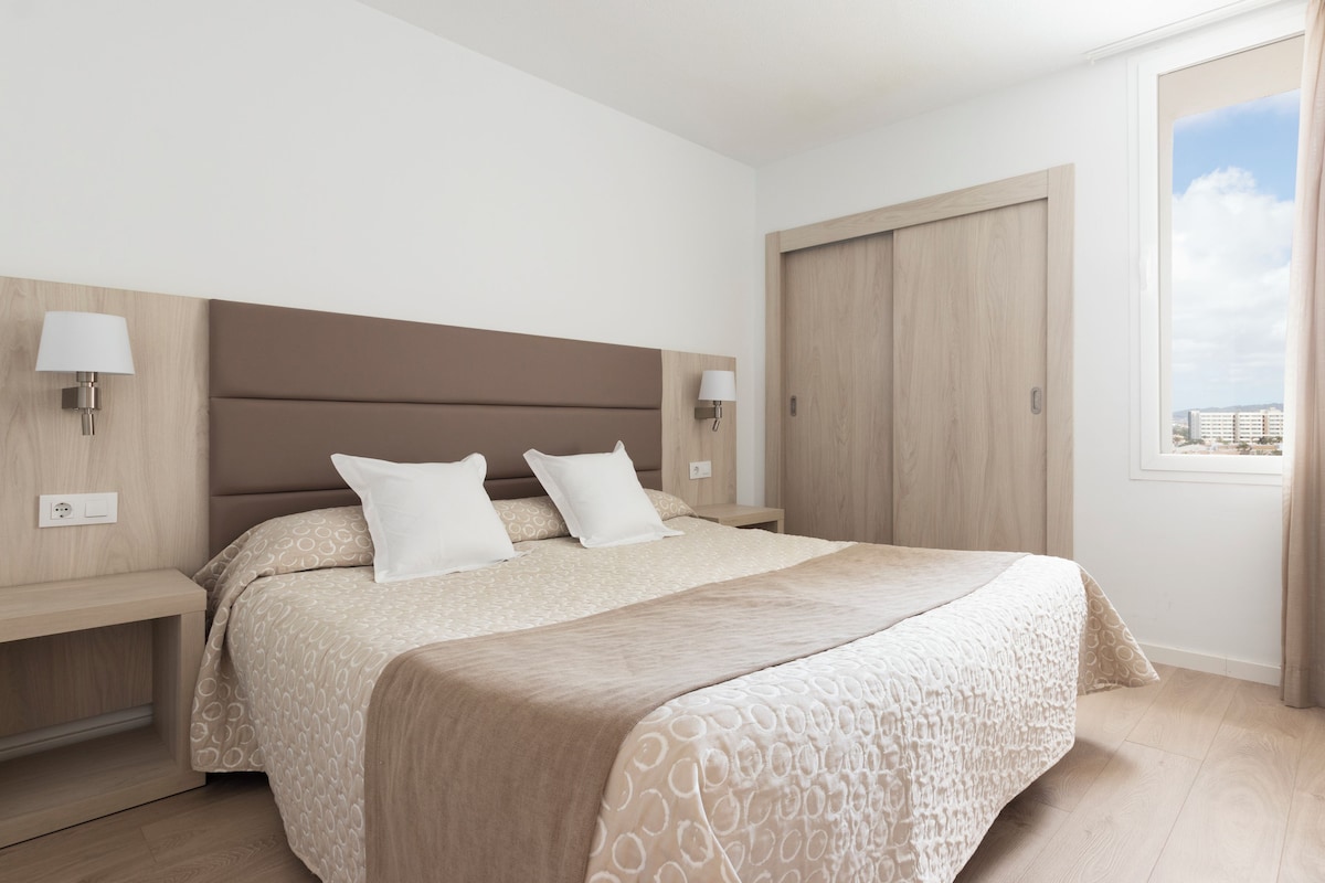 A modern hotel room with a large bed, two bedside tables, a nightstand, a wardrobe, and a window with curtains.