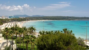 A beach with clear turquoise water, palm trees, and a hotel in the background.