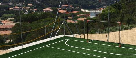 Una cancha de tenis al aire libre con red, rodeada por una valla, y vistas a un pueblo y montañas al fondo.