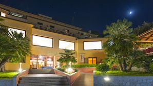 A hotel entrance with a staircase leading up to the doors, surrounded by well-maintained greenery and illuminated by ambient lighting.