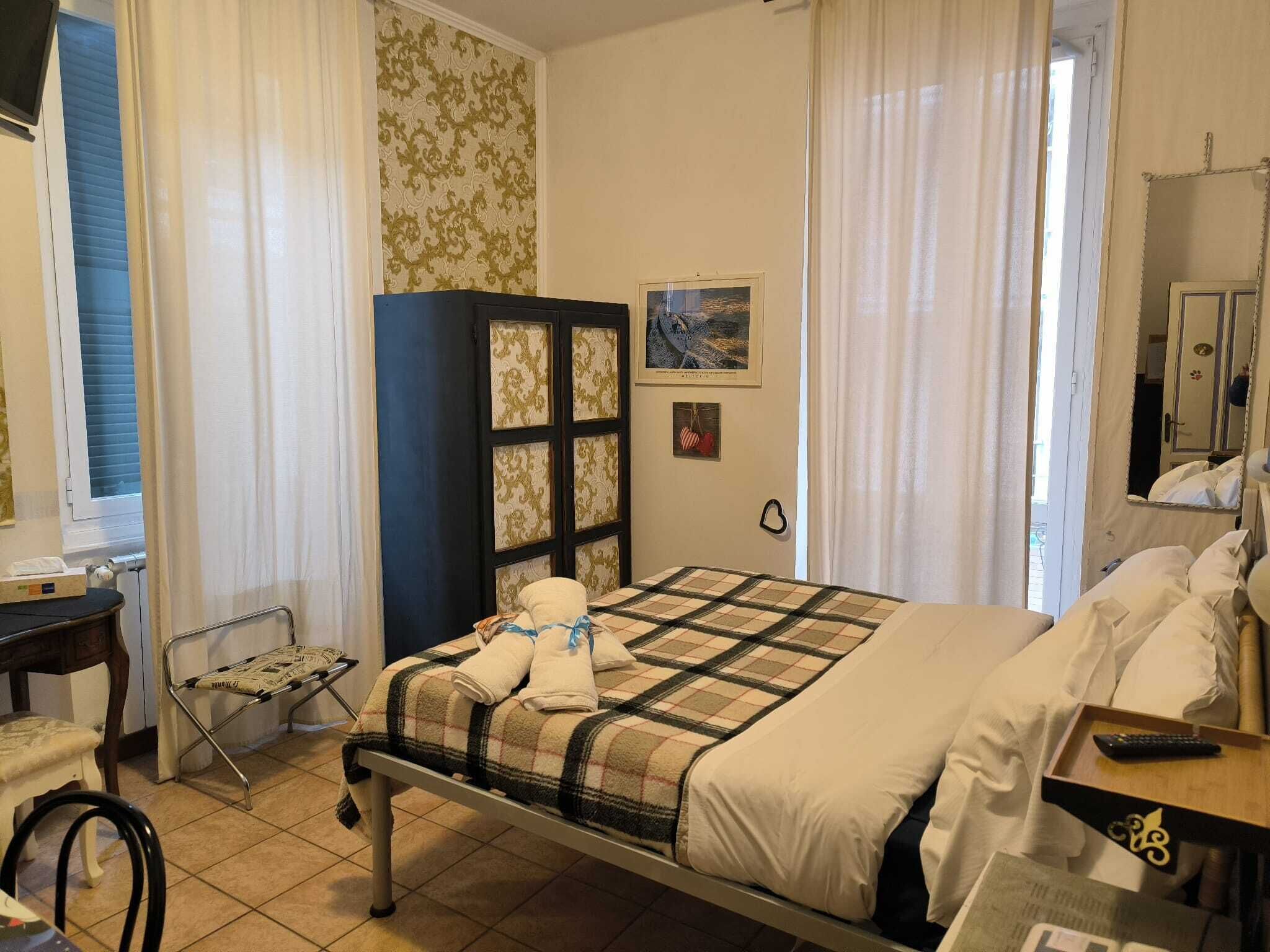 Ett hotellrum med en säng, en stol, ett litet bord, ett skåp och ett fönster med gardiner.