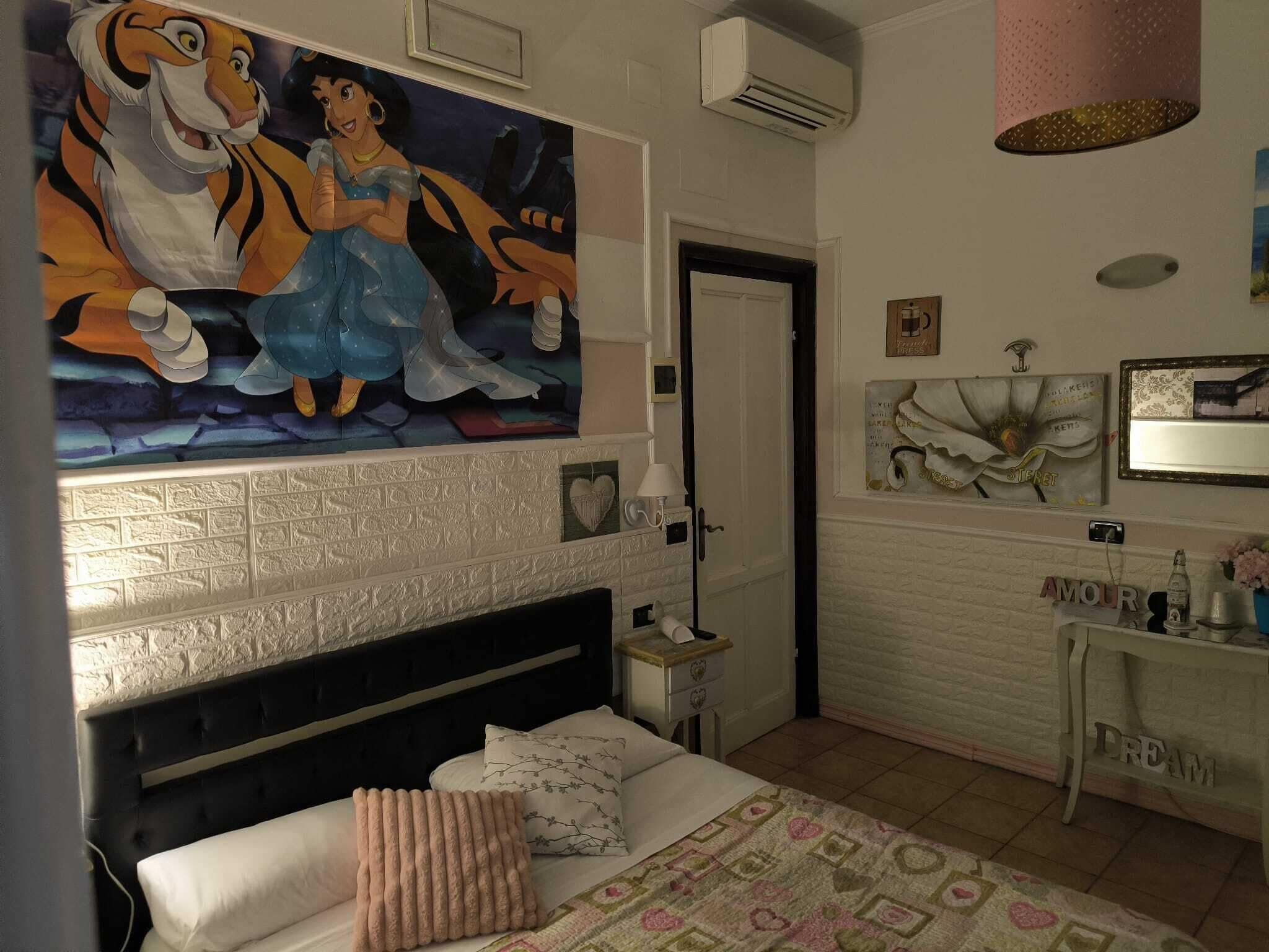 Een slaapkamer met een groot schilderij met Disney-thema, een wit bakstenen haardvuur, een bed met een quilt en een nachtkastje met een lamp.