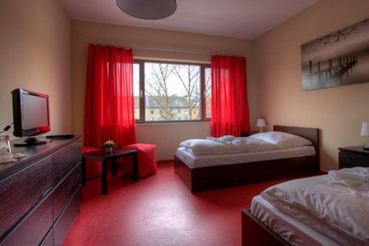 Une chambre à coucher avec un lit, un bureau avec un ordinateur, une fenêtre avec des rideaux rouges et un tableau accroché au mur.