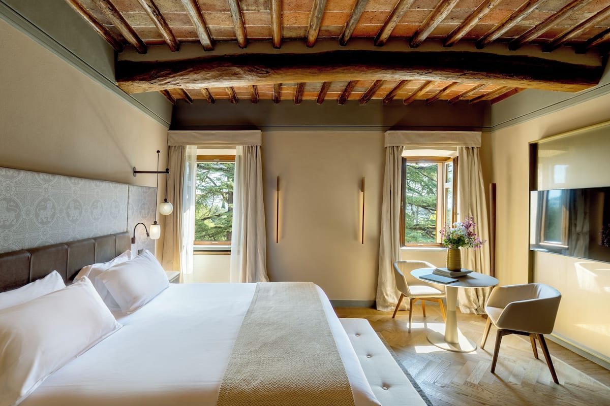 A hotel room with a large bed, a small table with a vase, and a chair. There are curtains on the windows and a wooden ceiling.