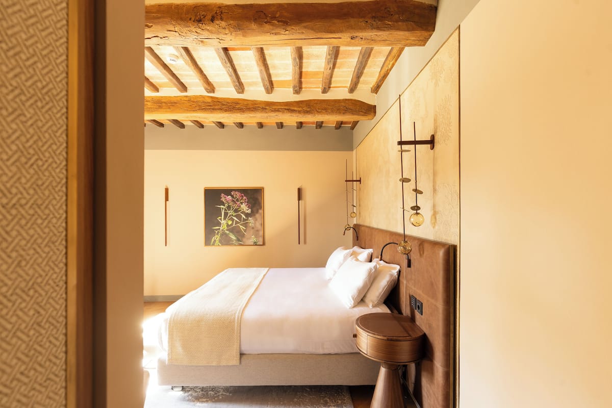 A bedroom with a wooden ceiling, a bed with white linens, a bedside table with a round mirror, and a wall-mounted light fixture.