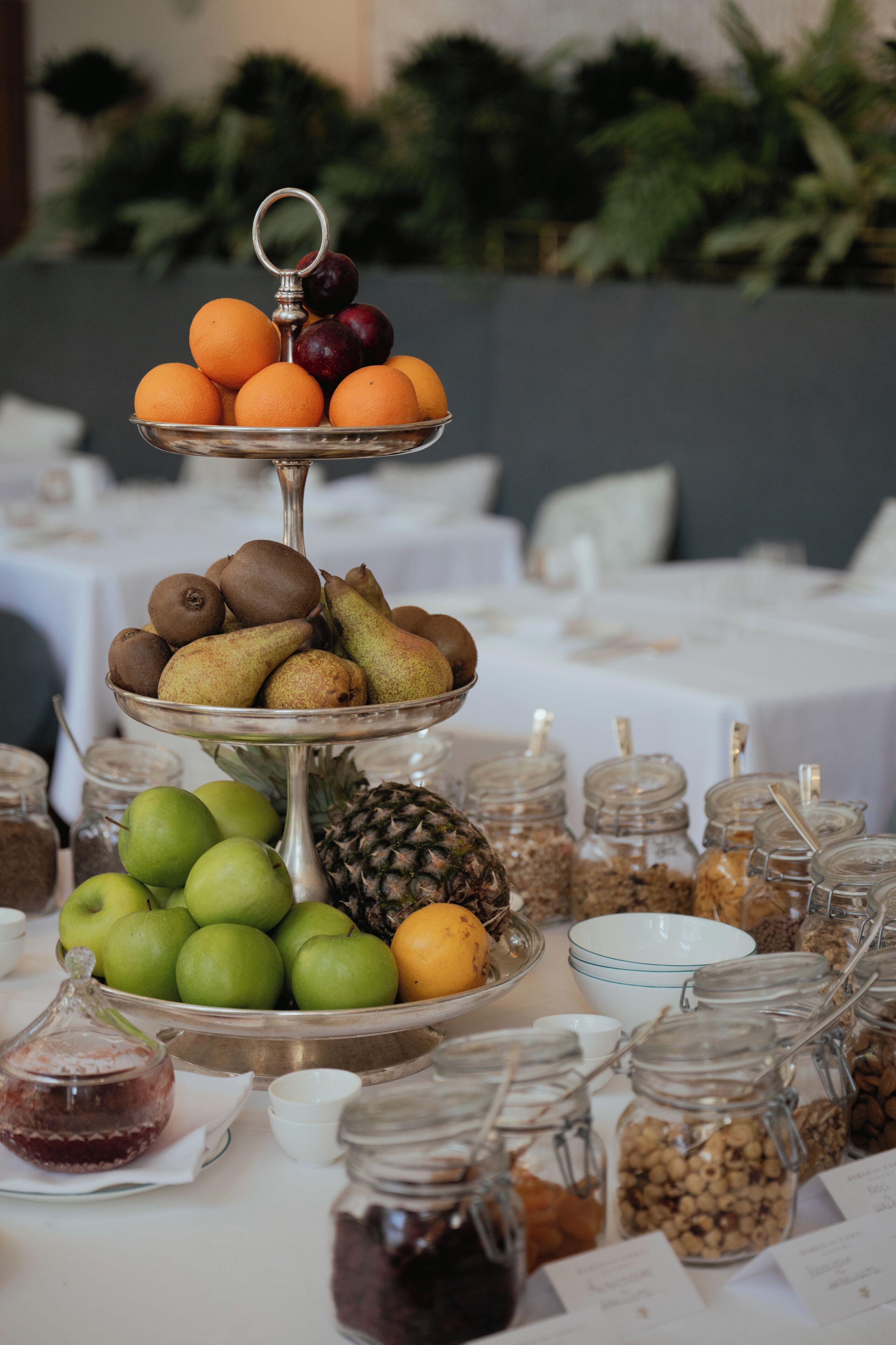 A multi-tiered fruit display with various fruits and jars of preserves on a table.