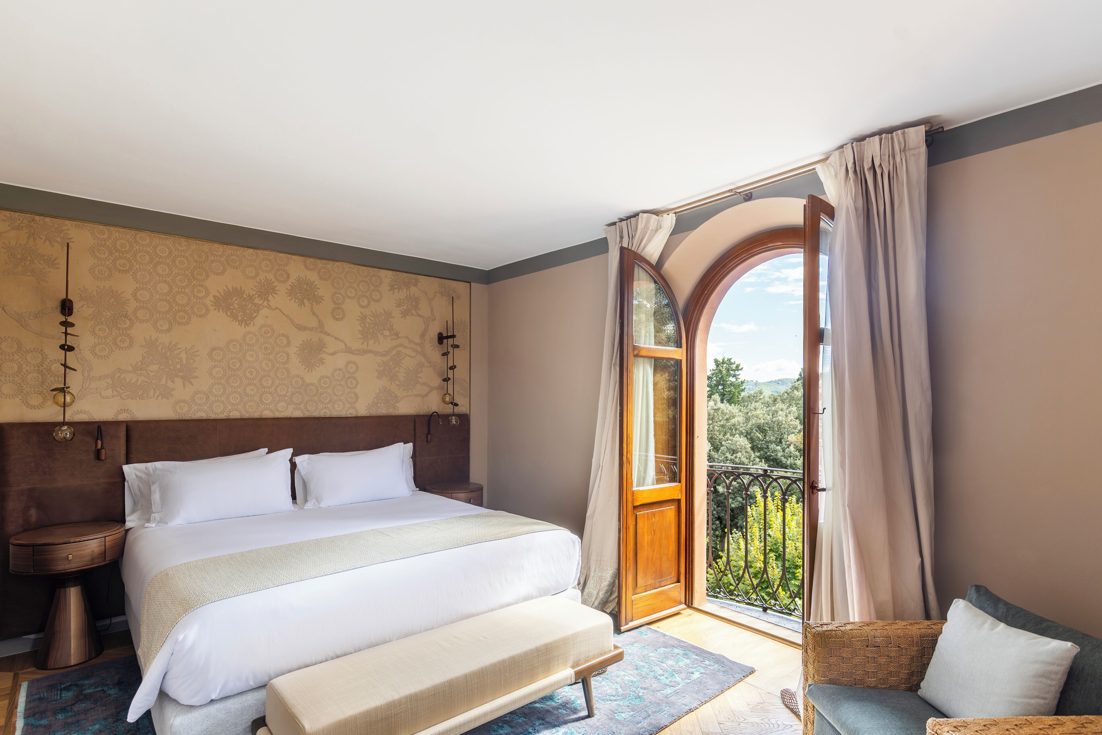 Ein Schlafzimmer mit einem großen Bett, einem Nachttisch, einer Bank, einem Korbstuhl und einem Balkon mit Vorhängen.