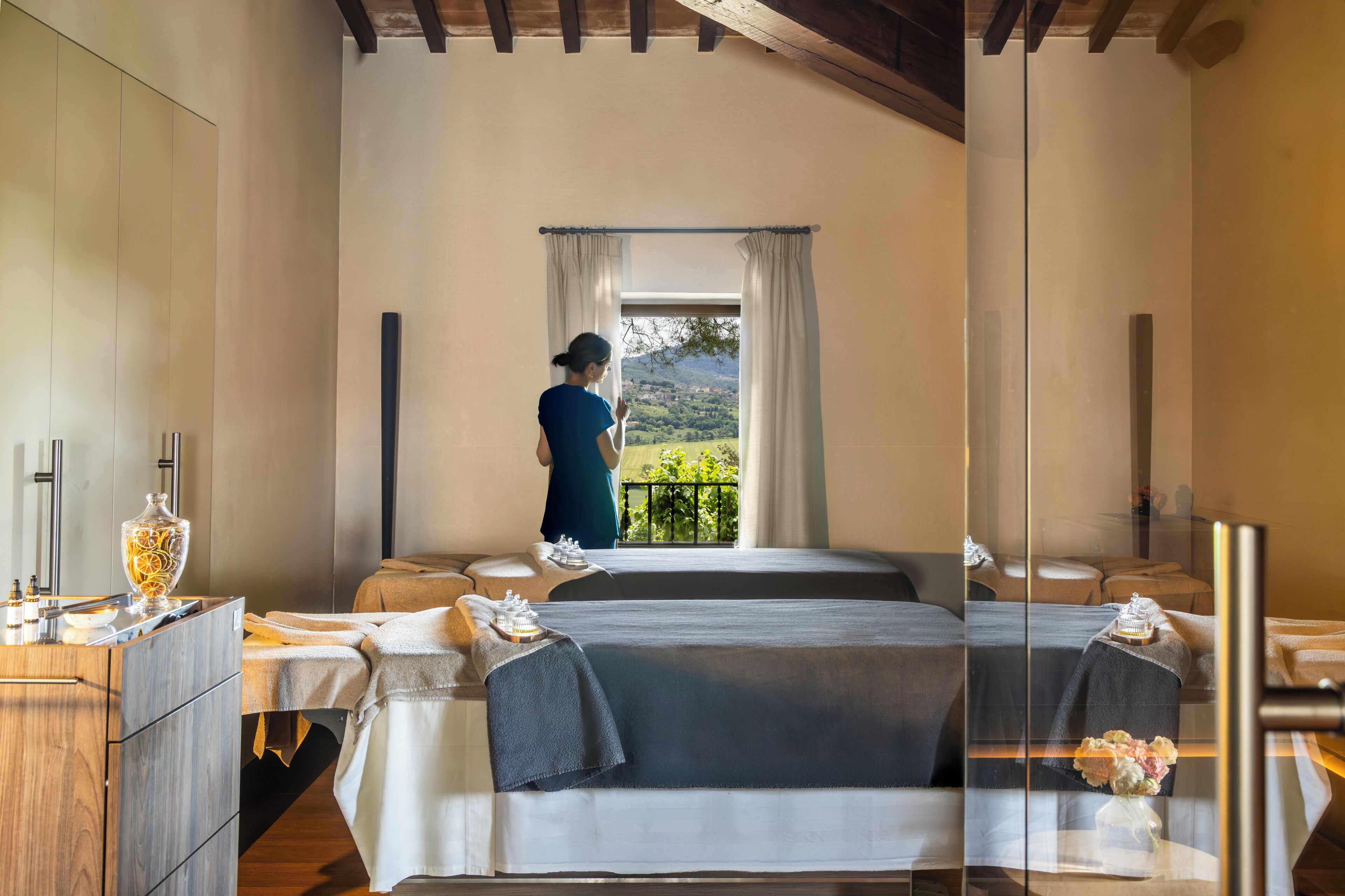 A room with two beds, a large window with curtains, a view of greenery outside, and a wooden ceiling.