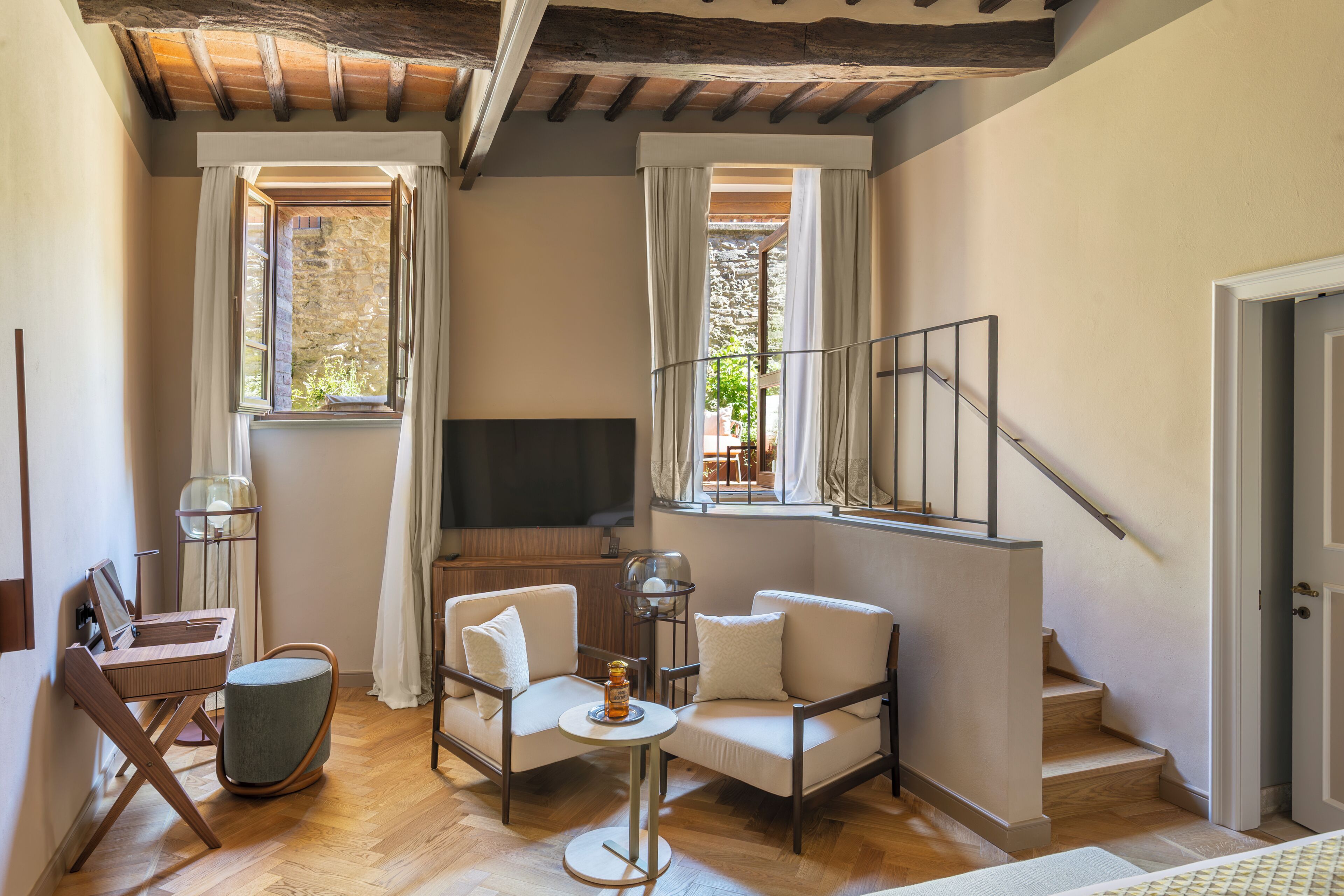 Un salon confortable avec un canapé, une petite table ronde et un téléviseur. Il y a deux fenêtres avec des rideaux, un plancher en bois et un escalier menant à un autre niveau.