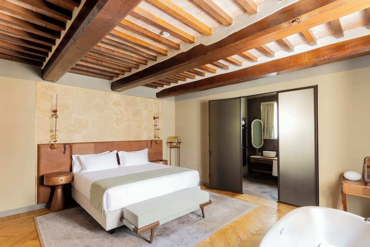 A bedroom with a large bed, wooden beams on the ceiling, a bathroom with a bathtub, and a mirror.