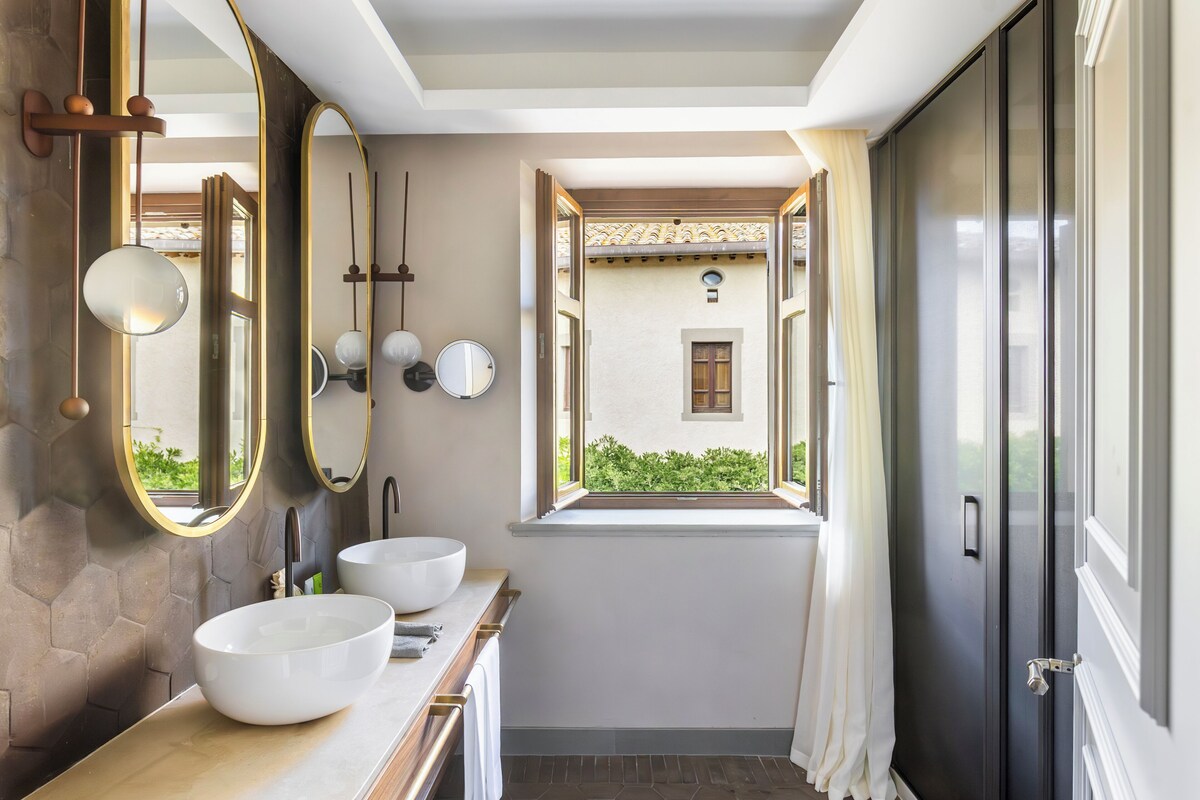A modern bathroom with two large white sinks, a mirror, and a window with a view of greenery.