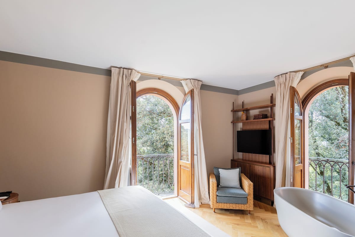 A bedroom with a large bed, a sitting area with a chair and a bathtub, and a balcony with a wooden railing.