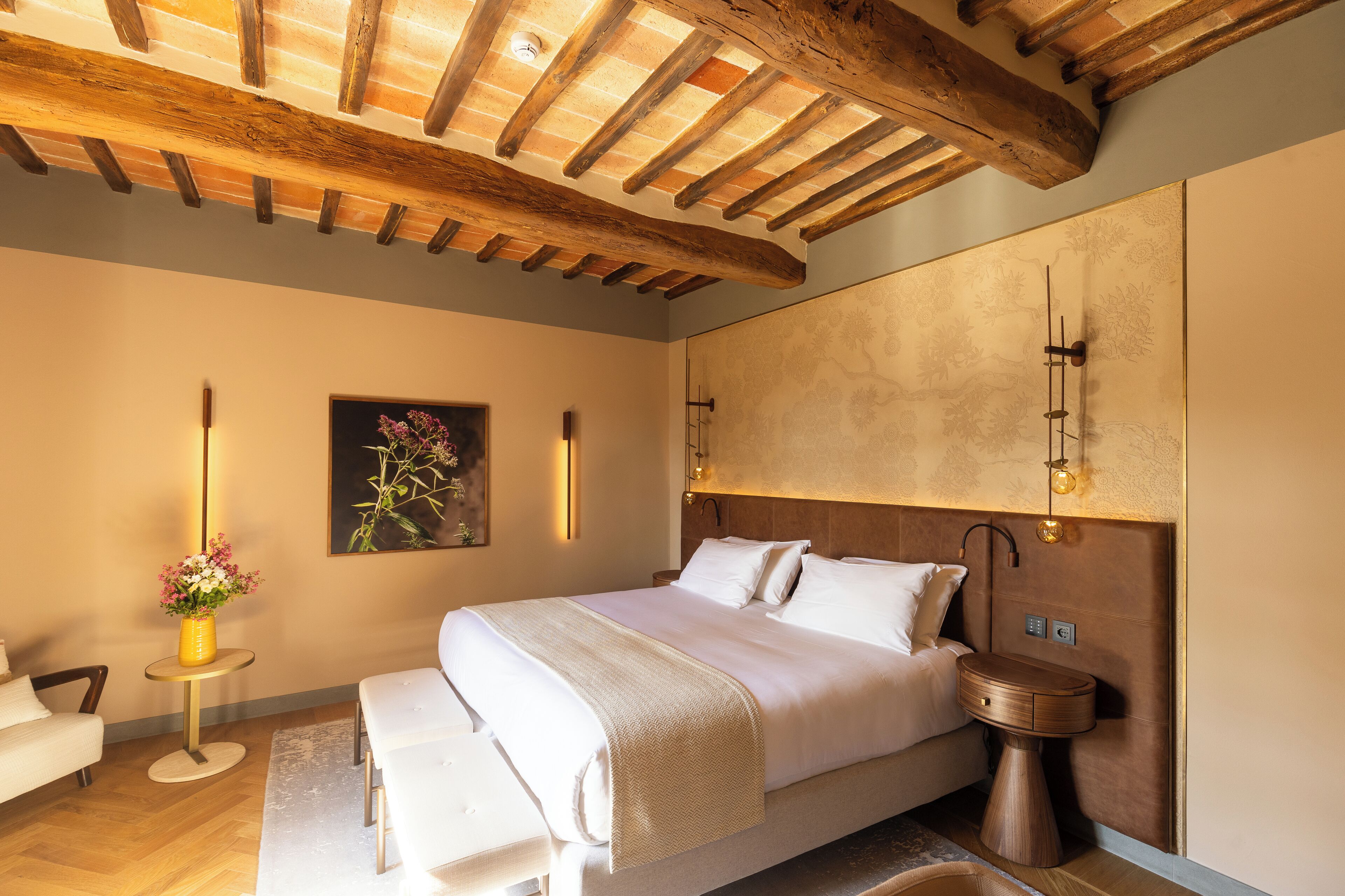 Un dormitorio con techo de madera, cama con ropa de cama blanca, mesita de noche con un jarrón de flores y una lámpara fijada a la pared.