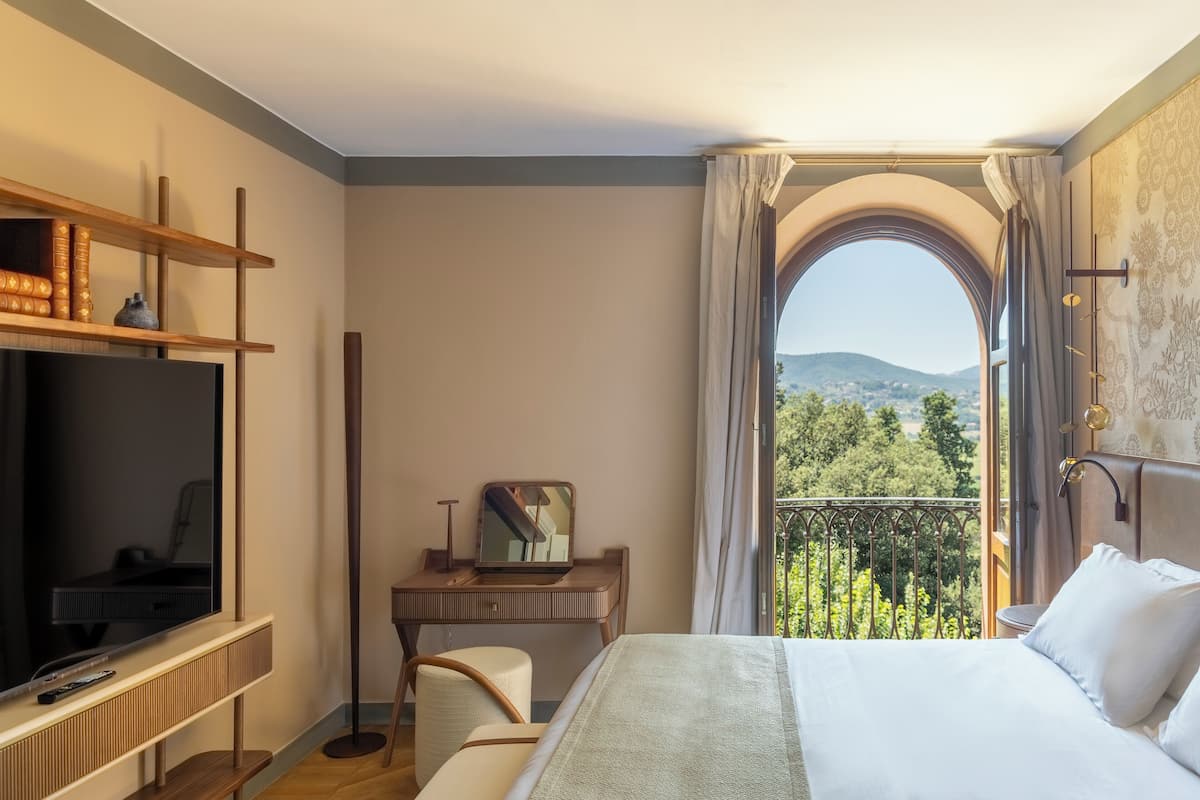 A hotel room with a bed, a desk with a computer, a TV, and a balcony with a view of the mountains.