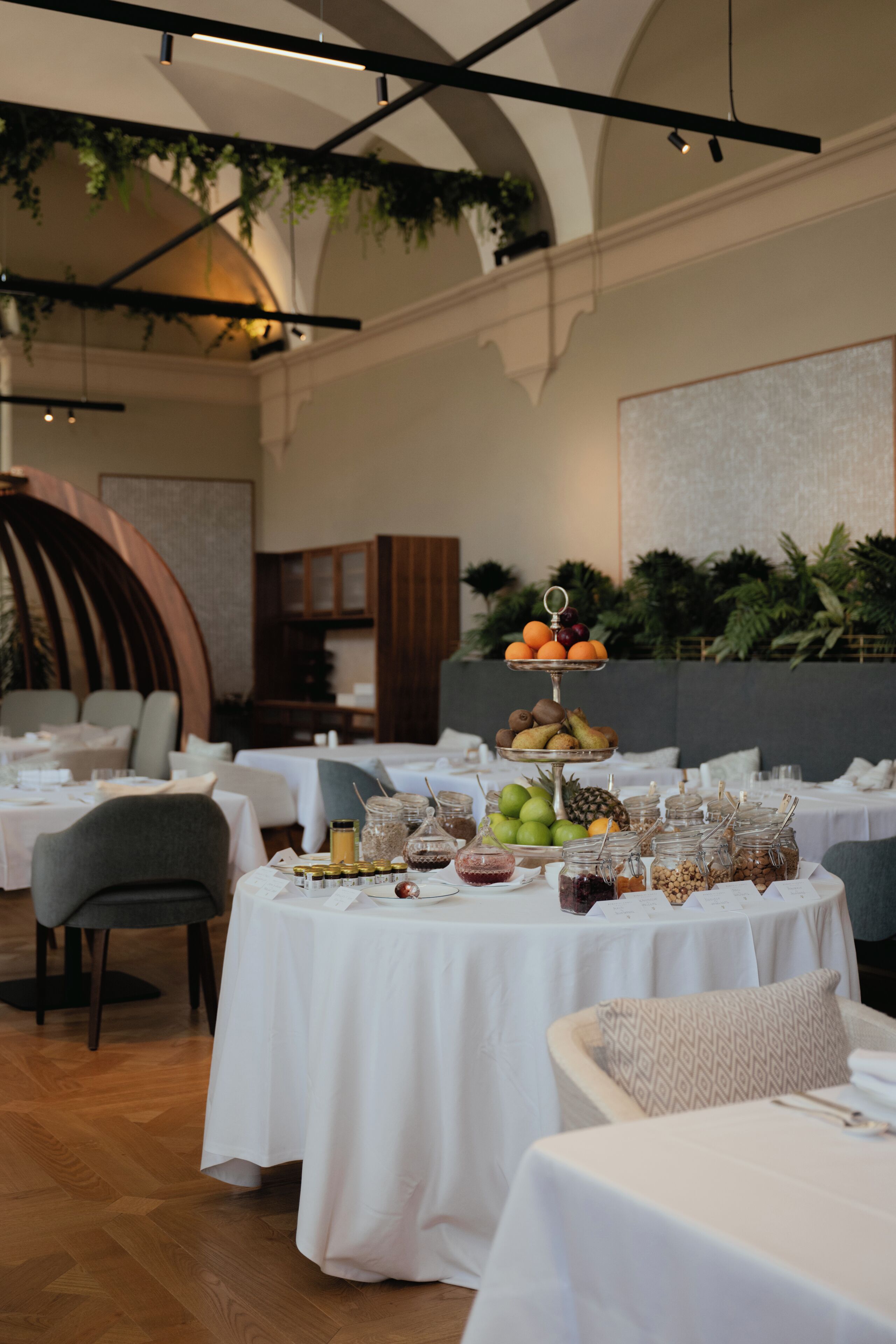 A restaurant interior with round tables covered in white tablecloths, a tiered fruit display, and various food and drink containers.