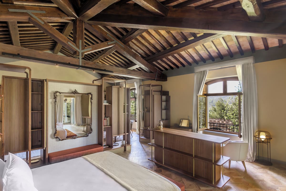 A bedroom with a large bed, wooden ceiling beams, a mirror, a desk, and a view of the outdoors.
