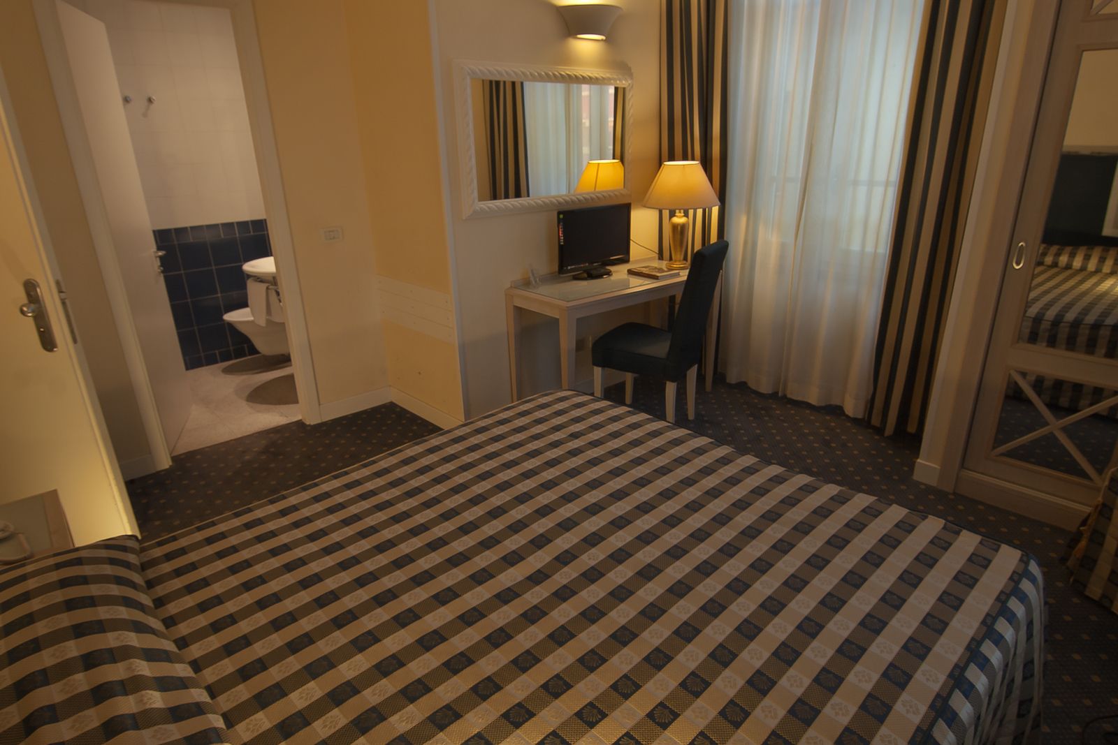 Una habitación de hotel con una colcha a cuadros, un escritorio con computadora, una silla, un espejo y una ventana con cortinas.