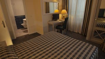 A hotel room with a checkered bedspread, a desk with a computer, a chair, a mirror, and a window with curtains.