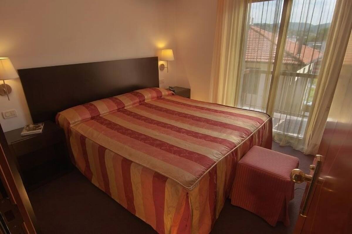 A hotel room with a large bed, a small table, and a view of the outside through a window.