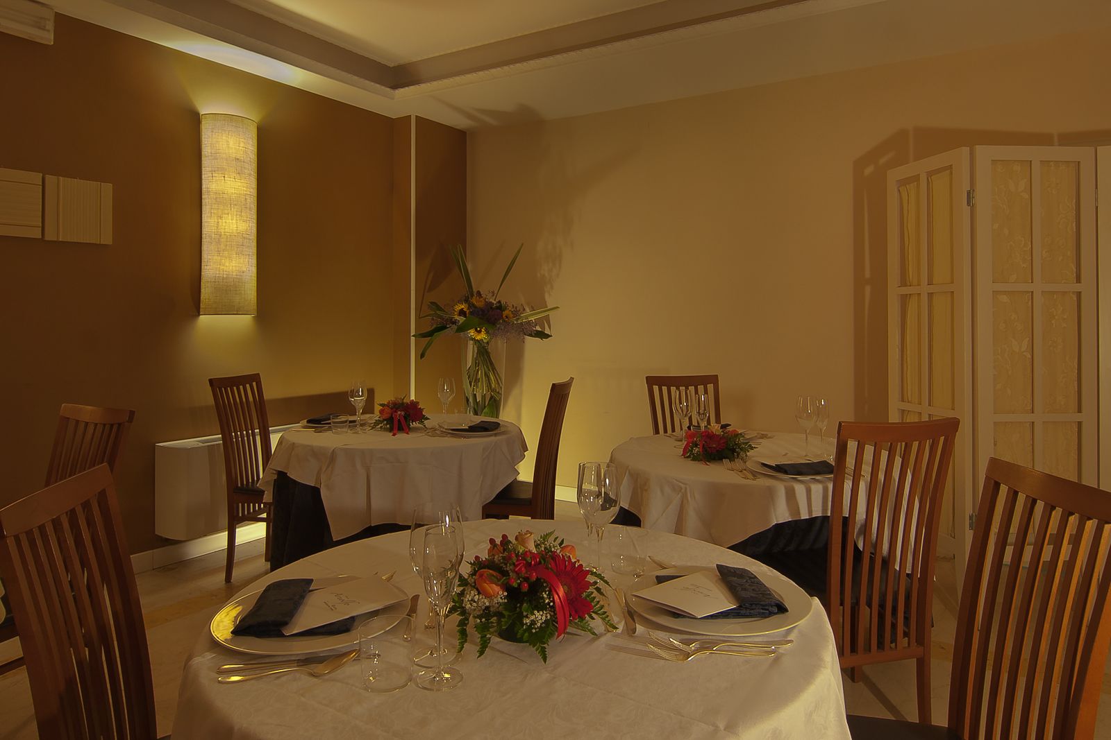 A dining area with round tables, each set with plates, cutlery, and glasses. There are chairs around the tables, and a vase with flowers on one table.
