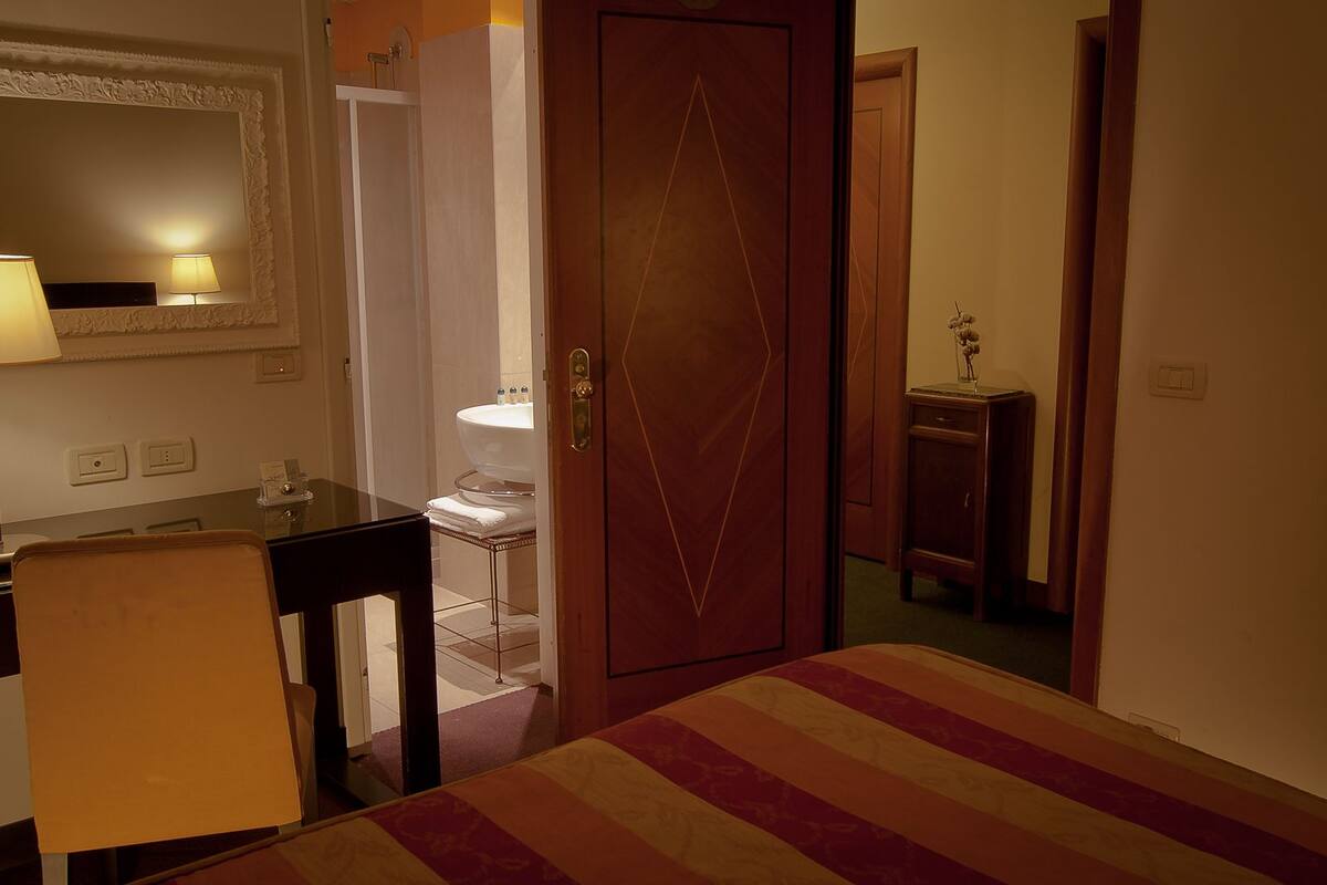 A hotel room with a bed, a desk, a chair, a bathroom visible through an open door, and a mirror on the wall.