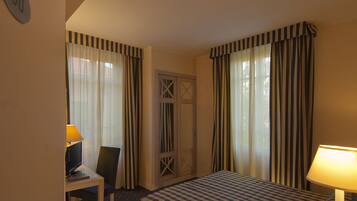 A hotel room with a bed, a desk with a chair, a lamp, and a door with a window.