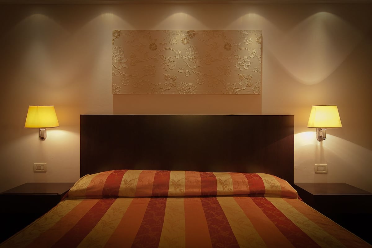 A hotel room with a large bed, two bedside lamps, a wall-mounted artwork, and a control panel on the wall.