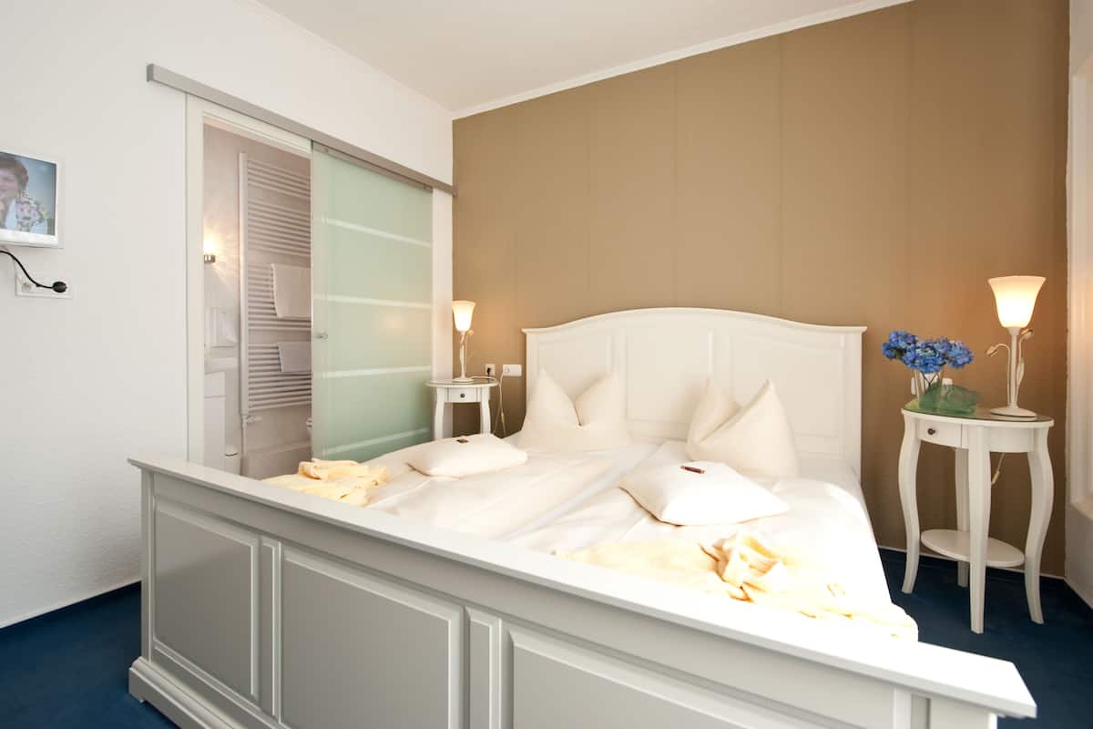 Una cama bien hecha con ropa blanca, dos mesitas de noche con lámparas y vista a un baño con tendedero.