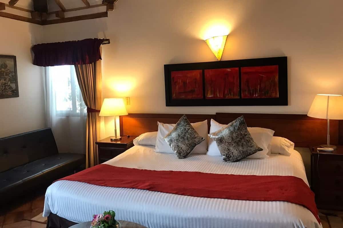 Una habitación de hotel con una cama grande, dos lámparas de noche, una manta roja y una obra de arte enmarcada sobre la cama.