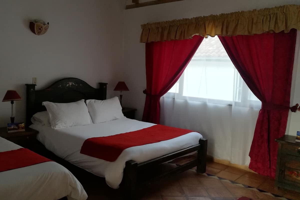 Cama doble con ropa de cama blanca y una manta roja, dos mesitas de noche con lámparas y una ventana con cortinas rojas.