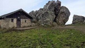 Um edifício rústico de pedra com telhado inclinado, localizado numa área gramada com grandes formações rochosas ao fundo.