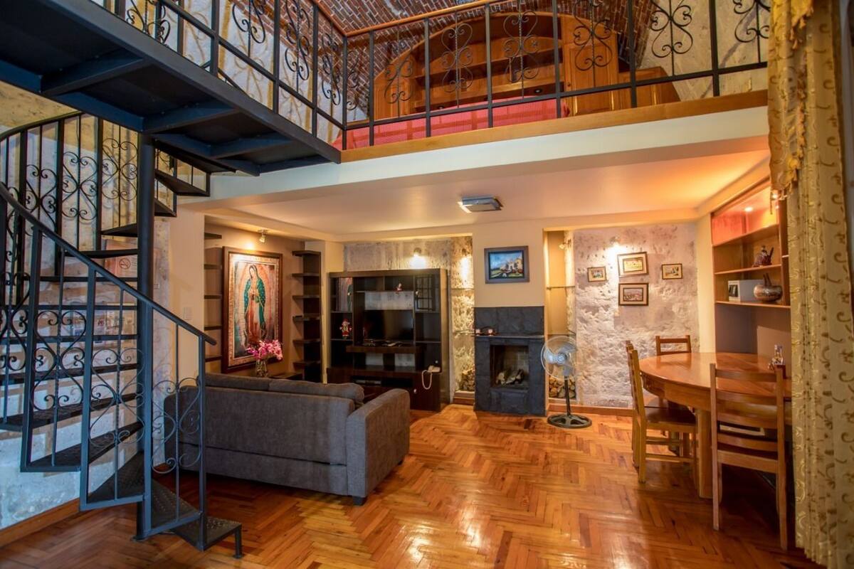 Un amplio salón con suelo de madera, chimenea, mesa de comedor y escalera con barandilla de hierro forjado.
