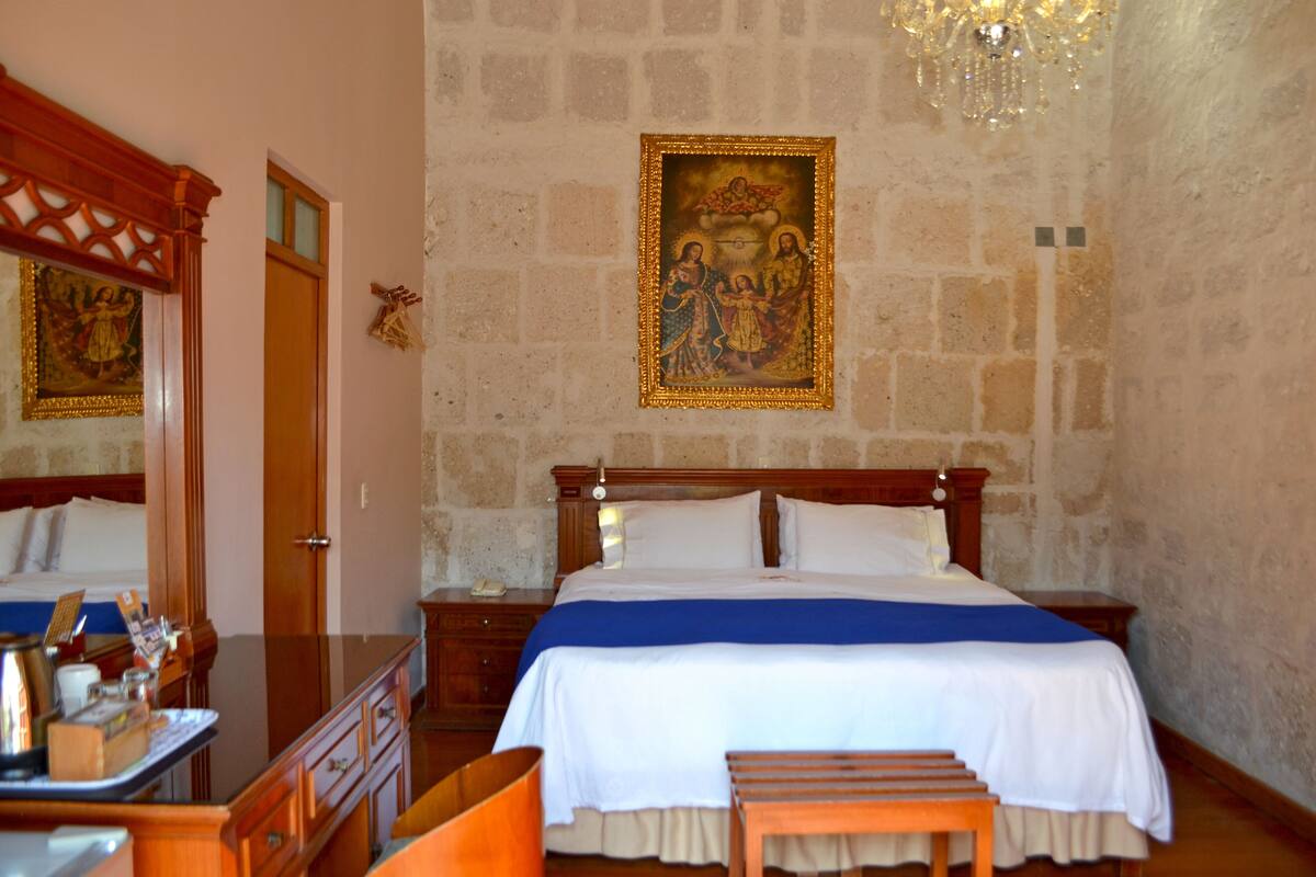 Habitación de hotel con una cama grande, muebles de madera, una araña y un cuadro religioso en la pared.