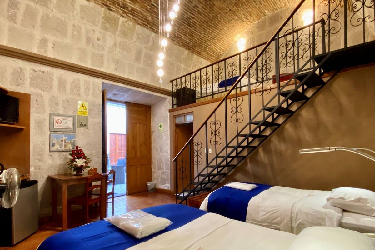 Una habitación con una cama individual, un escritorio con silla, una pequeña mesa con un florero y una escalera, además de un televisor fijado a la pared.