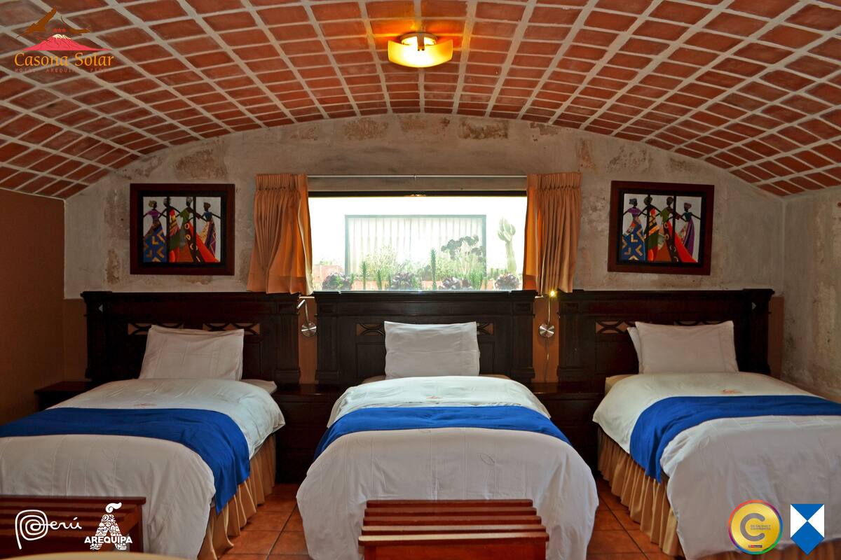 Habitación de hotel con tres camas, cabecera de madera, ventana con cortinas y obras de arte en las paredes.