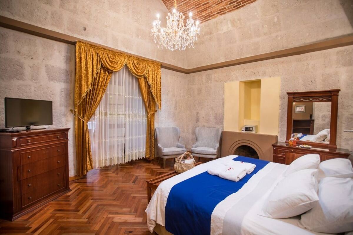 Una habitación de hotel con piso de madera en diseño espiga, cama con ropa de cama blanca y azul, televisor sobre un aparador de madera, dos sillones, un candelabro y un espejo con chimenea.