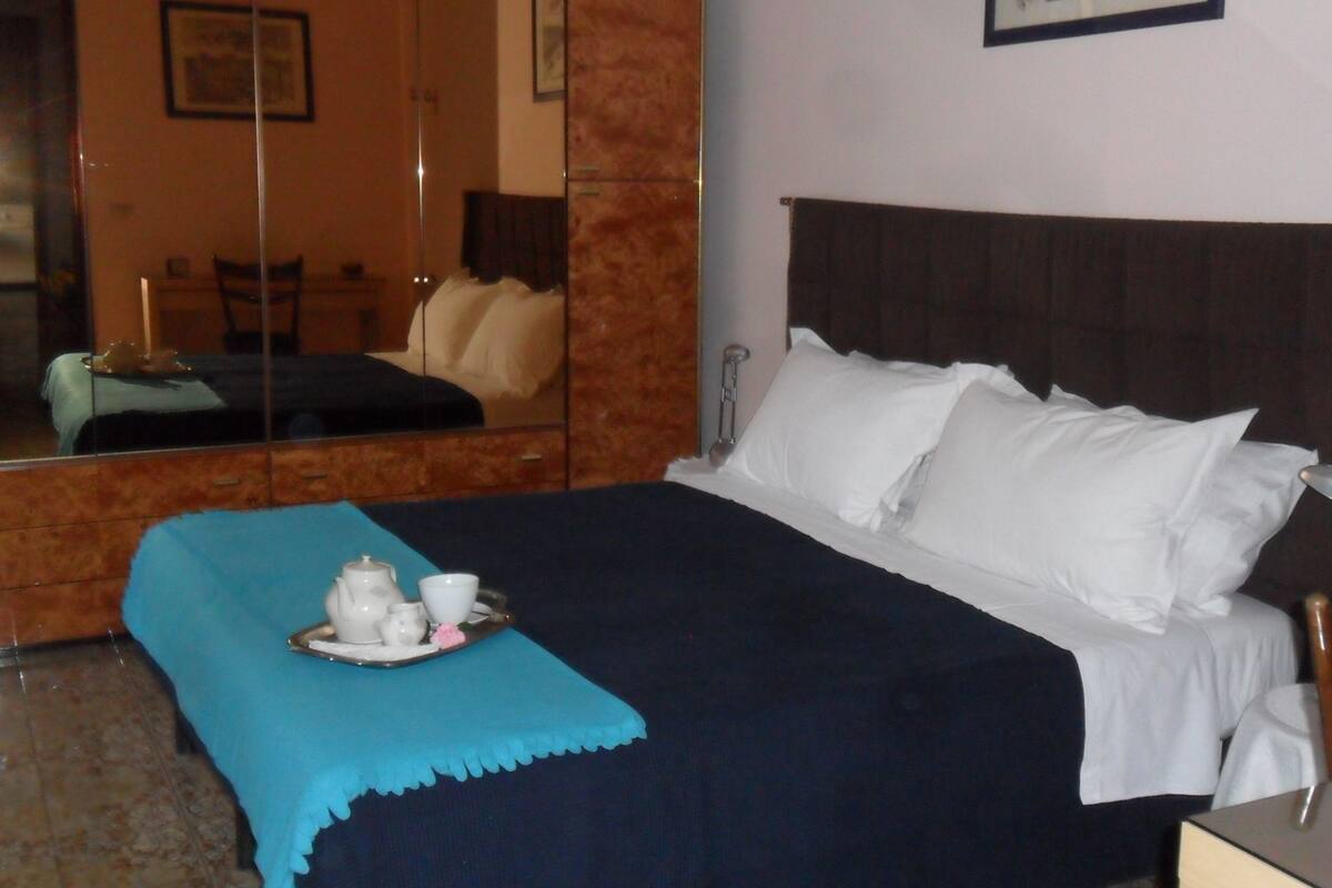 A neatly made bed with white linens and a blue throw blanket. A tray with tea cups and a teapot is placed on the bed.