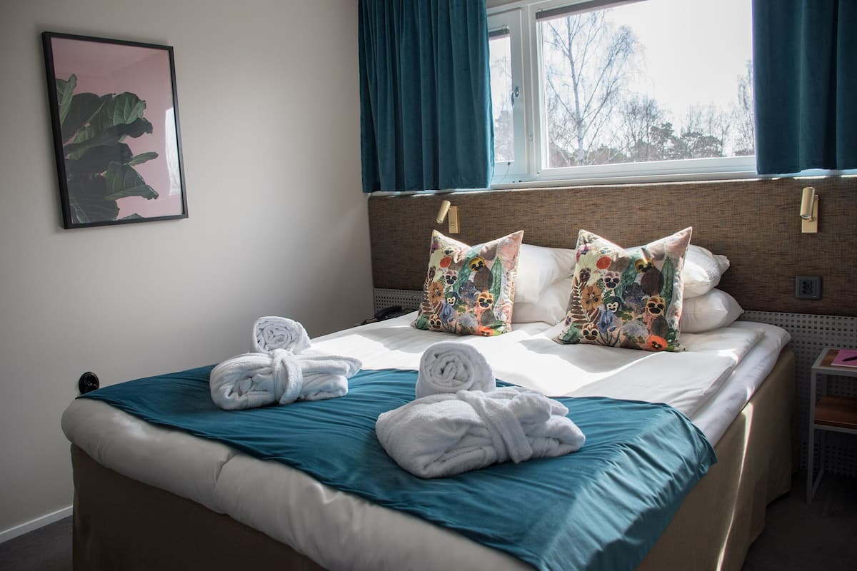 Una cama bien hecha con una colcha color aguamarina, sábanas blancas y almohadas decorativas. Se aprecia un cuadro en la pared y una ventana con cortinas.