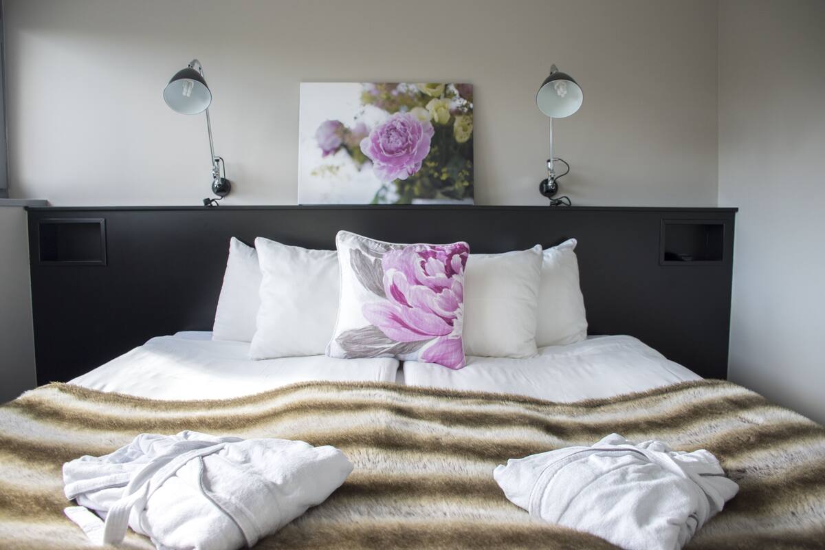 Una cama bien hecha con sábanas blancas y una manta a rayas, dos lámparas de noche y una obra de arte floral en la pared.