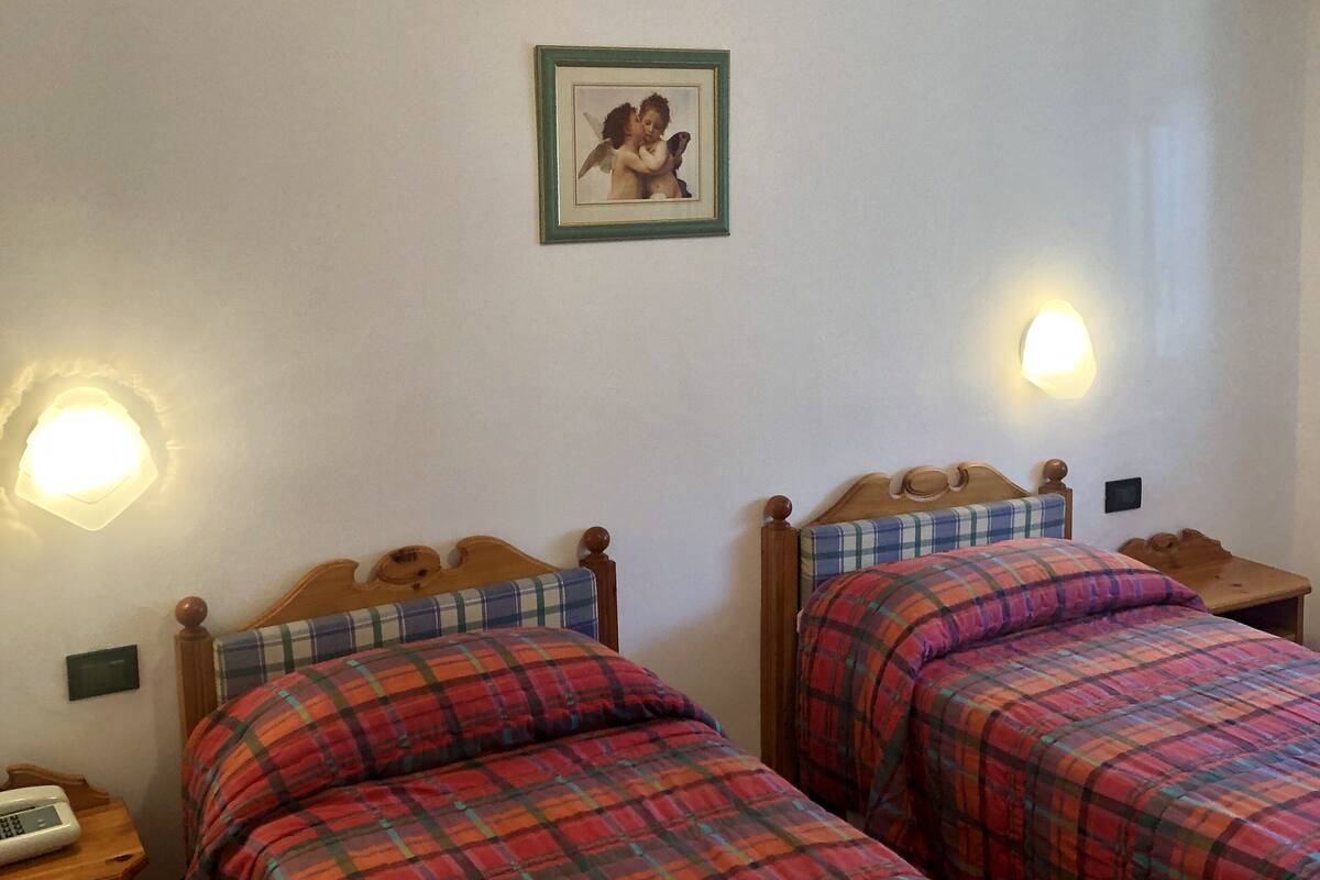 A room with two wooden beds, each with a plaid bedspread. There are bedside tables with lamps and a framed picture on the wall.