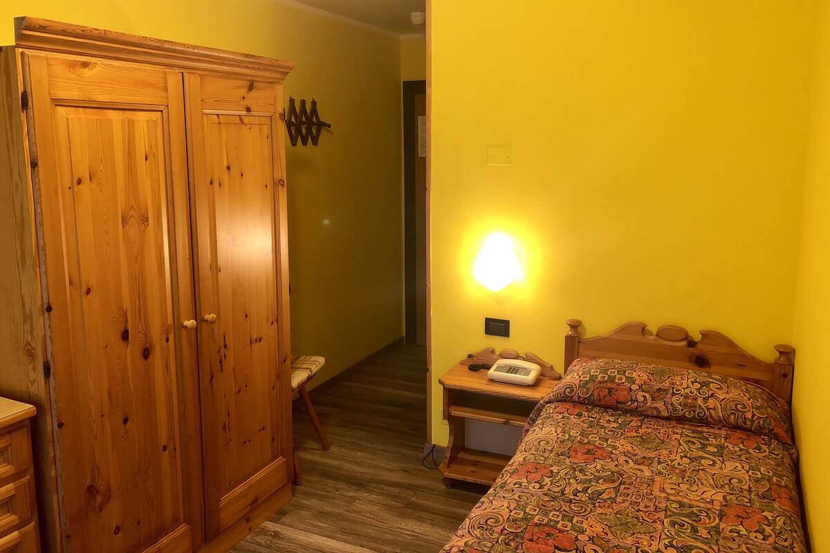 A room with a wooden wardrobe, a bed with a patterned bedspread, a bedside table with a lamp, and a chair.